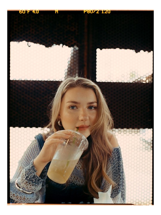 A woman sips a drink through a straw from a plastic cup, looking directly at the viewer.