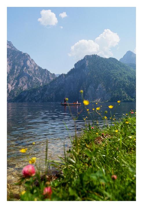 A small boat with two people floats on a lake surrounded by mountains, with wildflowers and grass in the foreground.