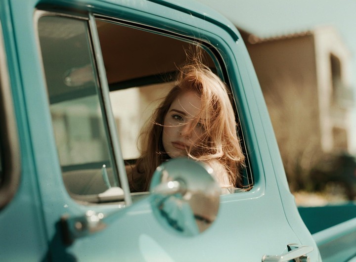 A woman with long red hair looks out the window of a light blue truck.