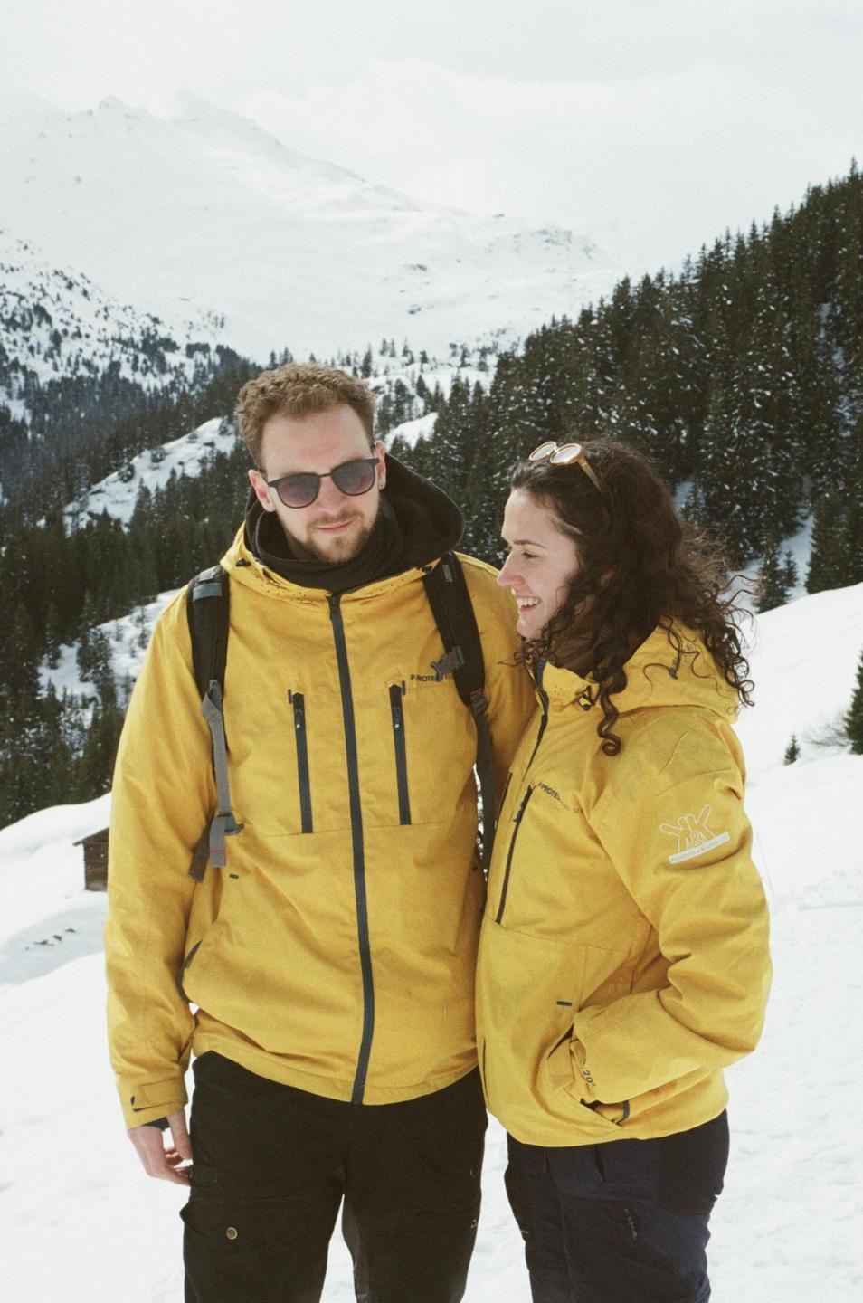 A couple wearing yellow jackets stands in a snowy mountain landscape with pine trees.