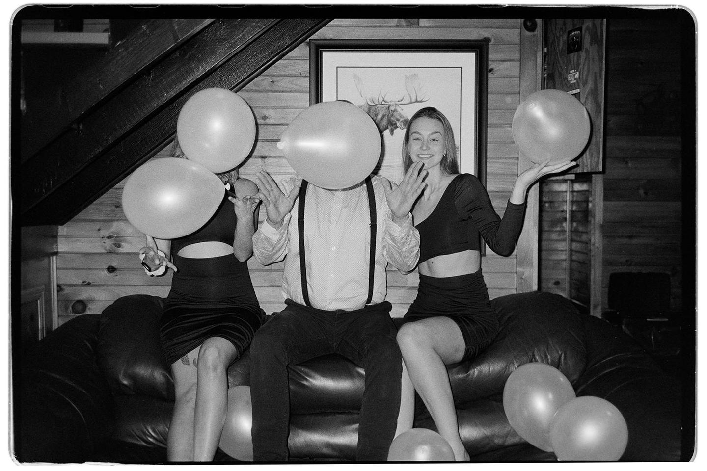 Three people sit on a couch with balloons, one of whom has a balloon covering their face.