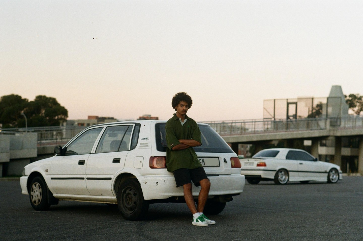 A man with curly hair leans against the back of a white car in a parking lot with another white car and an overpass in the background.