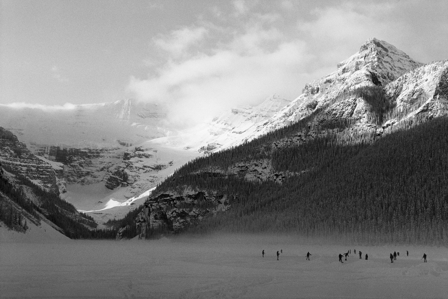 A group of people are ice skating on a frozen lake surrounded by snow-covered mountains and a forest.
