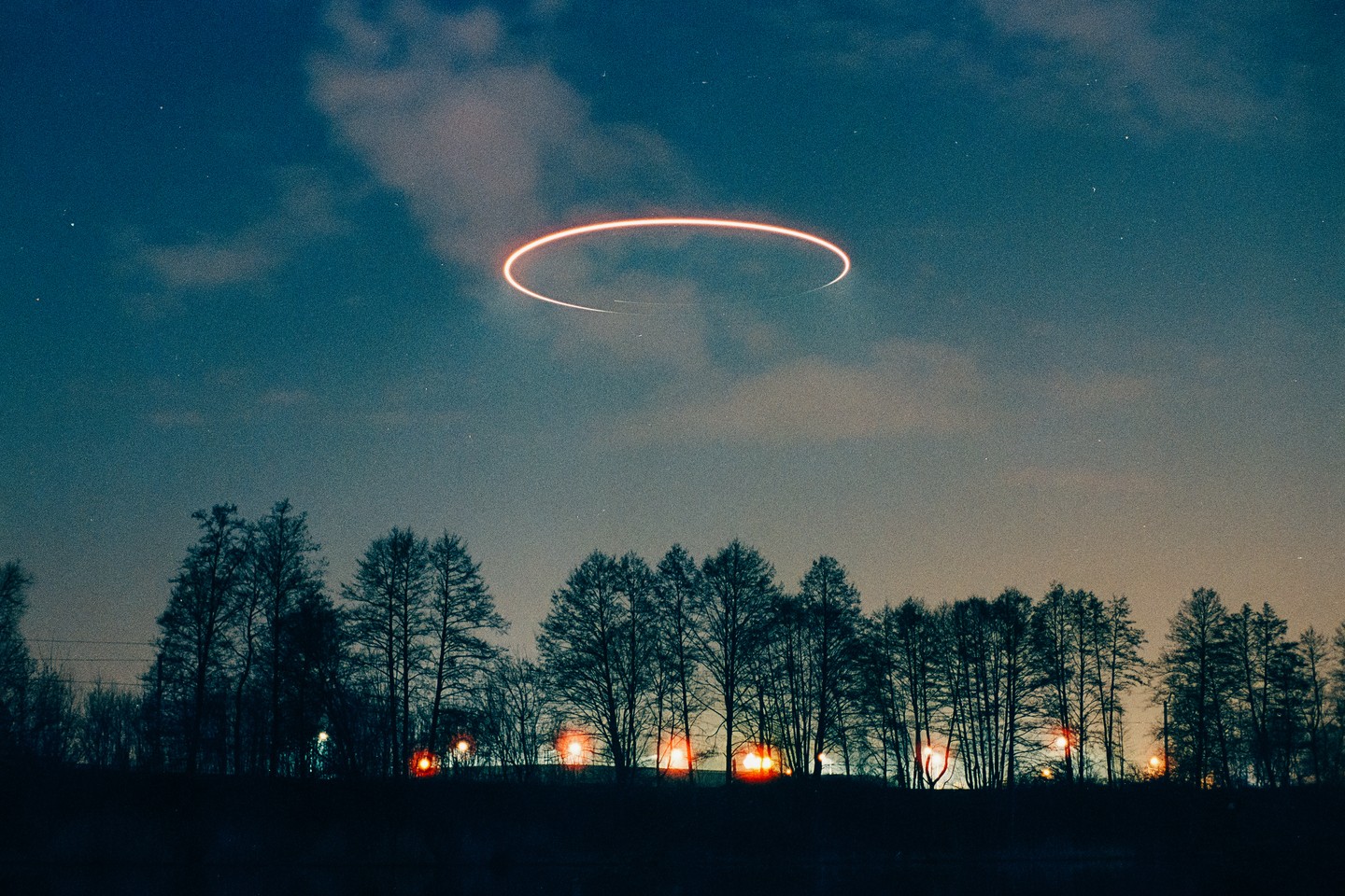 A glowing orange ring hovers in the night sky above a line of silhouetted trees with distant lights.