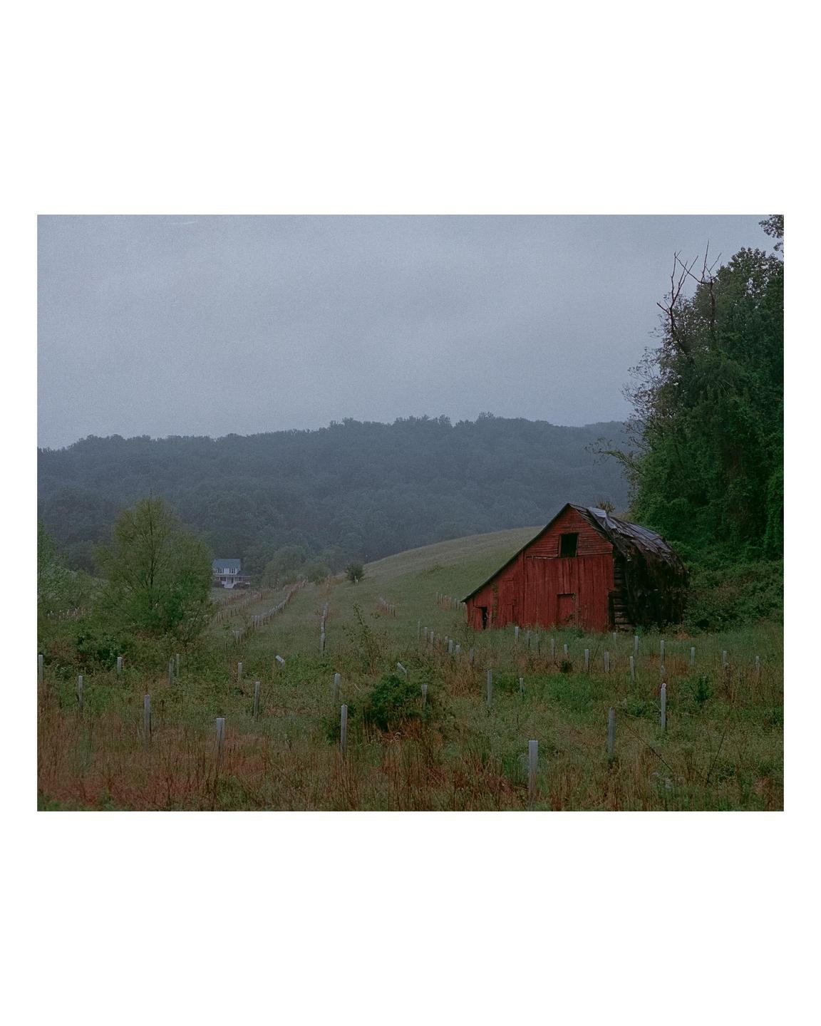 An old red barn sits on a grassy hill with a forest and hazy sky in the background.