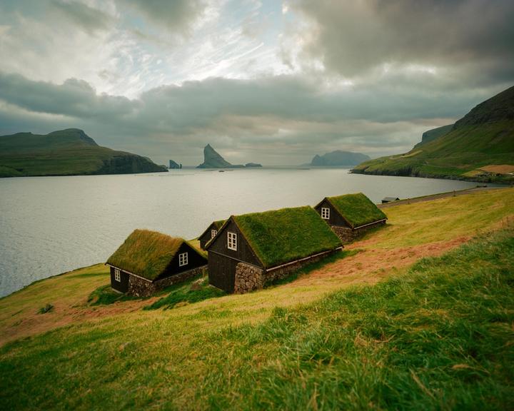 Three houses with grass roofs sit on a grassy hillside overlooking a body of water with mountains in the distance under a cloudy sky.