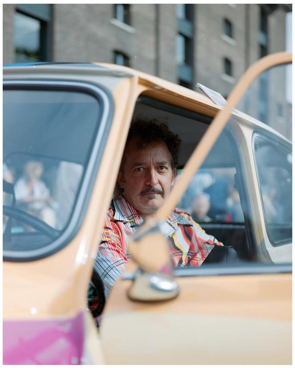 A man with a mustache and a colorful striped shirt sits in a beige car with the door open.