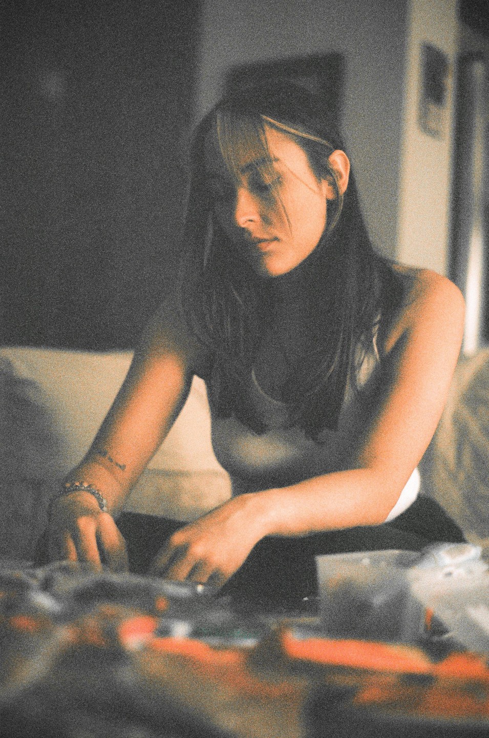 A woman with dark hair and a white tank top sits indoors, looking down at something in front of her.