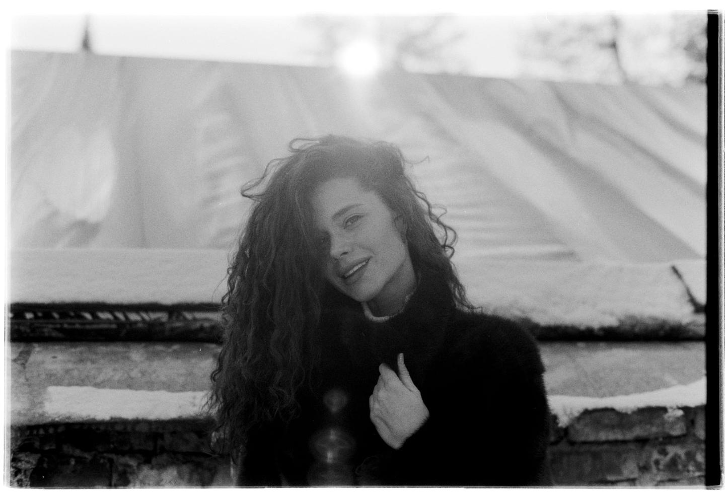 A woman with curly hair smiles at the viewer, with snow-covered surfaces and a bright light source in the background.