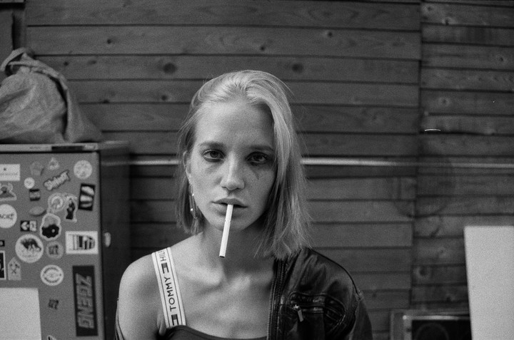 A woman with smudged eye makeup holds a cigarette in her mouth, wearing a leather jacket and a top with a branded strap, against a wooden wall and a sticker-covered surface.