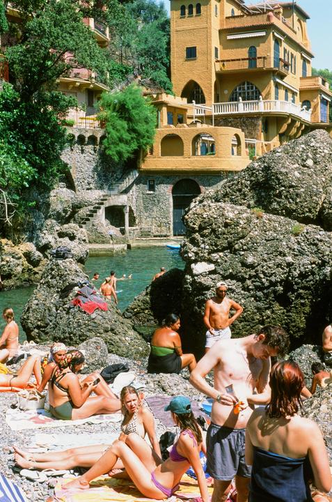 A group of people relax on a rocky beach next to a body of water with buildings on a hillside in the background.