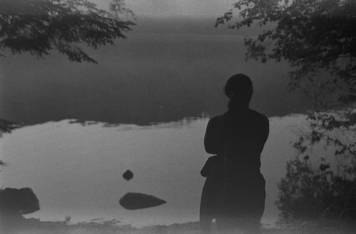 A person stands silhouetted against a body of water with trees framing the top of the image.