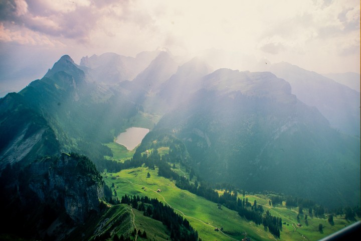 A high-angle landscape shows a mountain range with a lake in a valley, and a bright sky with clouds.