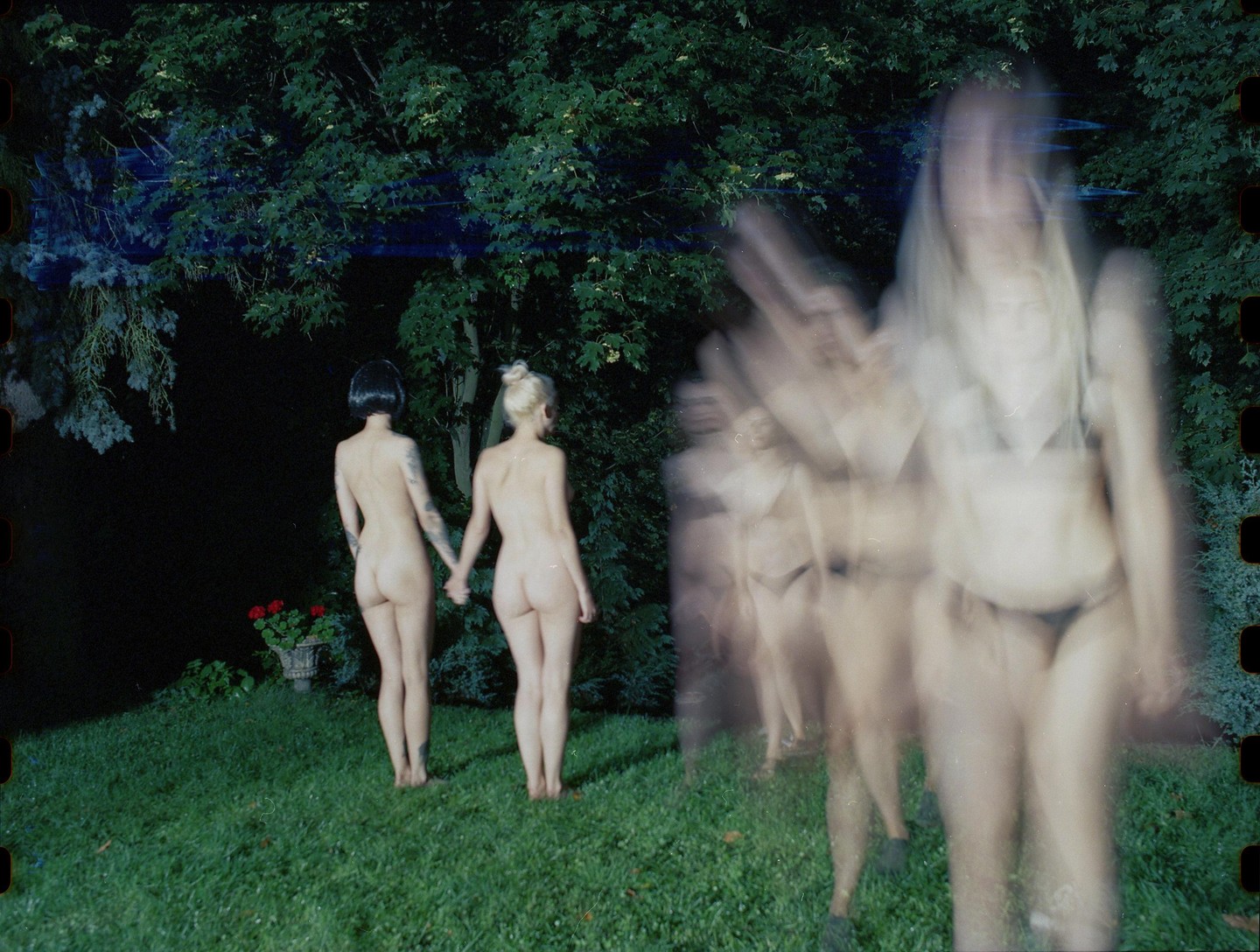 Two nude women hold hands in a garden at night while a blurred figure walks in the foreground.