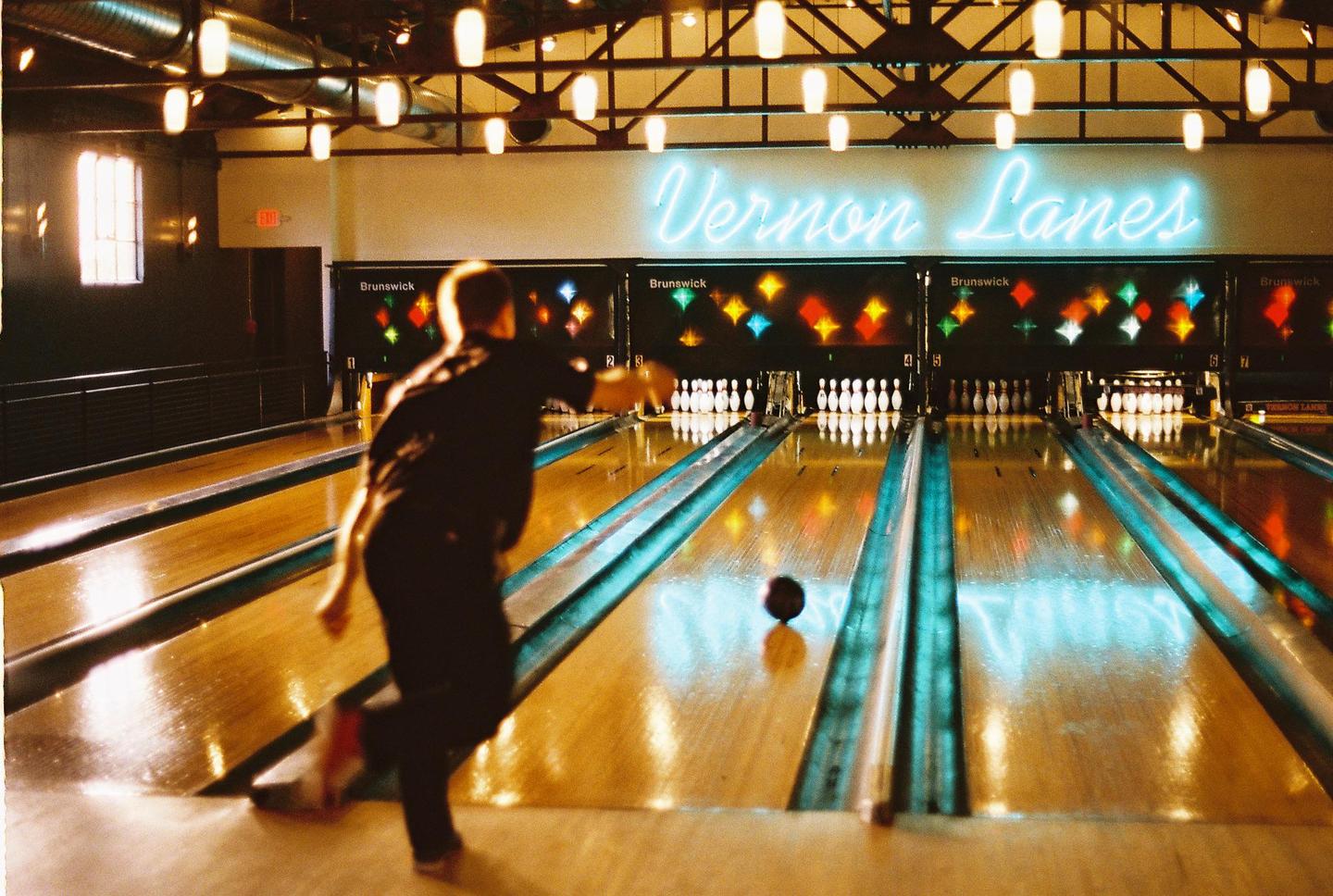 A man bowls a black bowling ball down a lane toward pins under a neon sign that reads "Vernon Lanes."