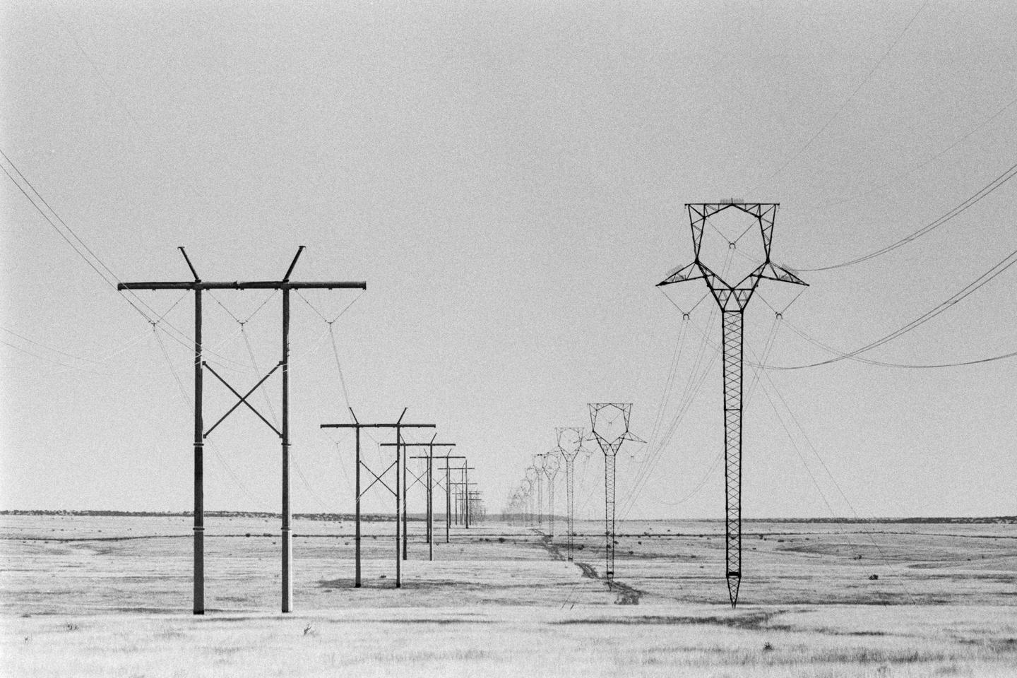 A line of power poles and transmission towers stretches into the distance across a flat, grassy landscape under a clear sky.