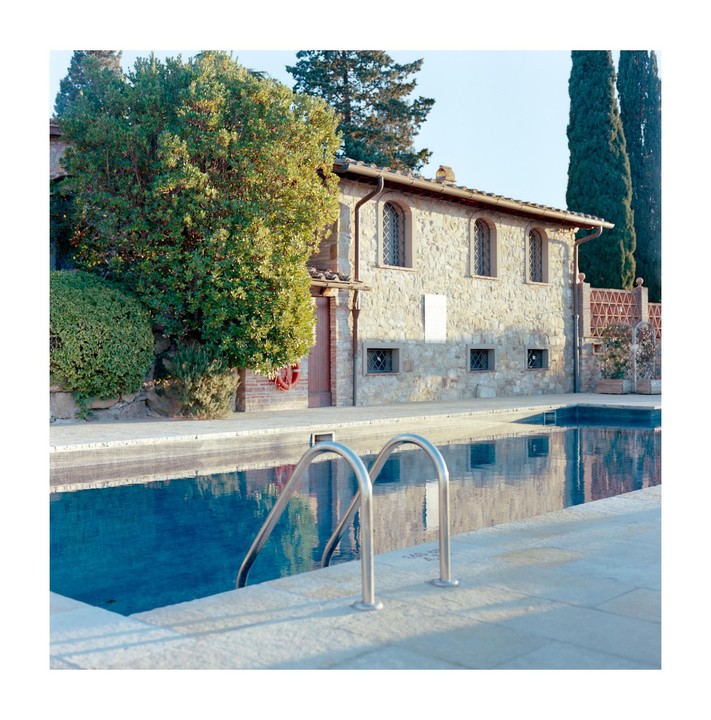 A stone building with arched windows stands beside a swimming pool with metal handrails under a clear sky.