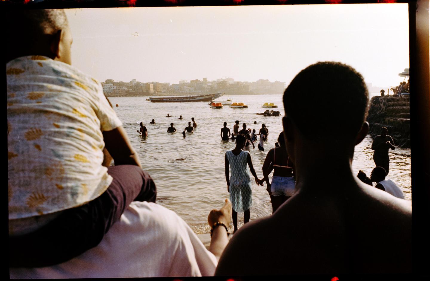 A child sits on a man's shoulders looking out at a body of water where many people are swimming and boats are docked.