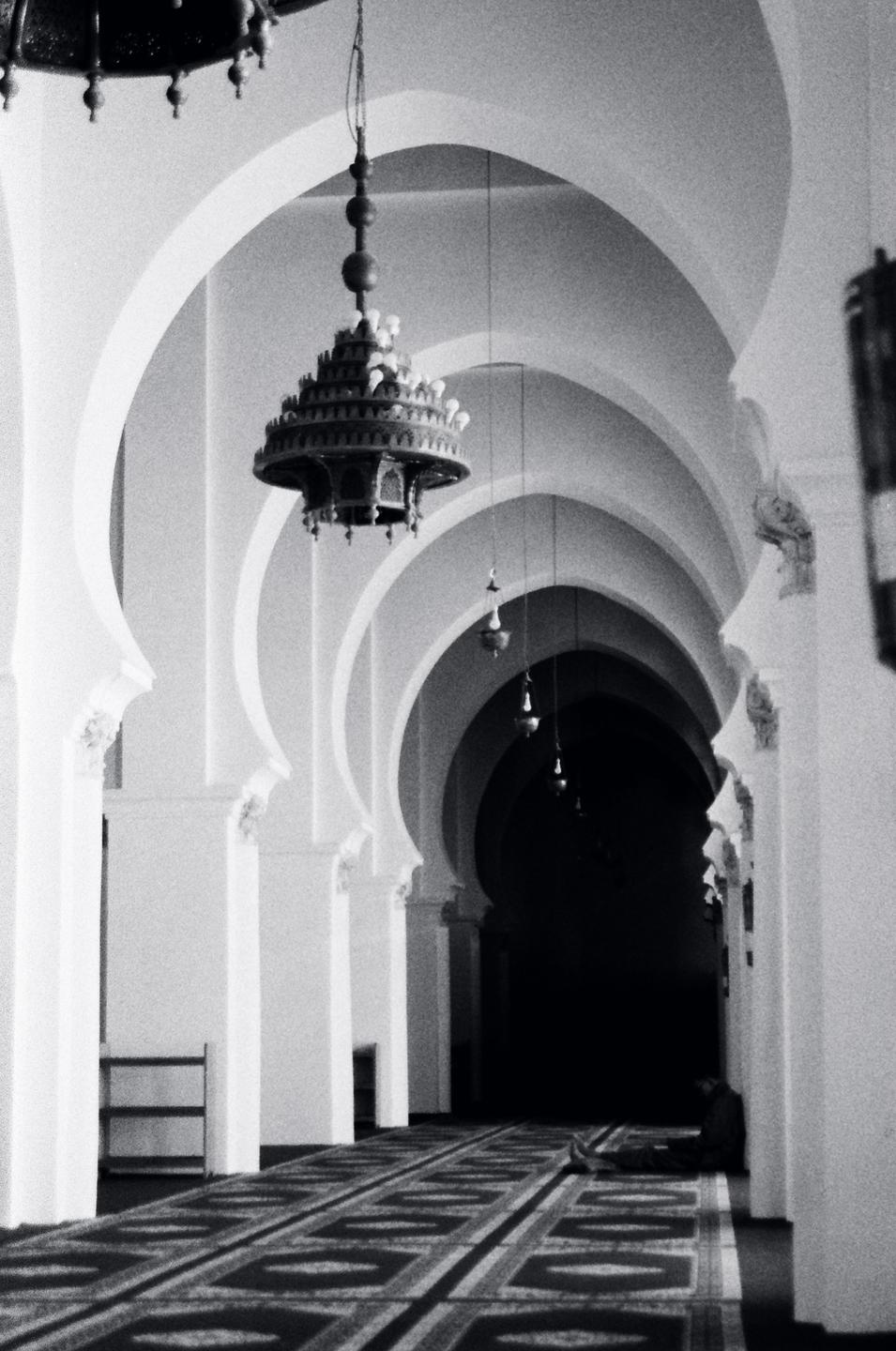 A person sits on a patterned carpet in a long hallway with arched columns and hanging lamps.