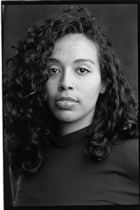 A woman with curly hair and nose and lip piercings looks directly at the viewer.