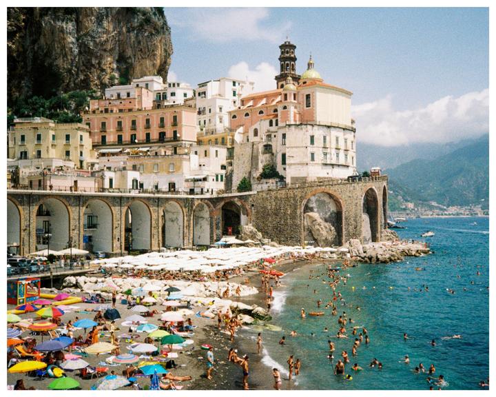 A crowded beach with many colorful umbrellas is next to a town built into a cliffside overlooking the sea.