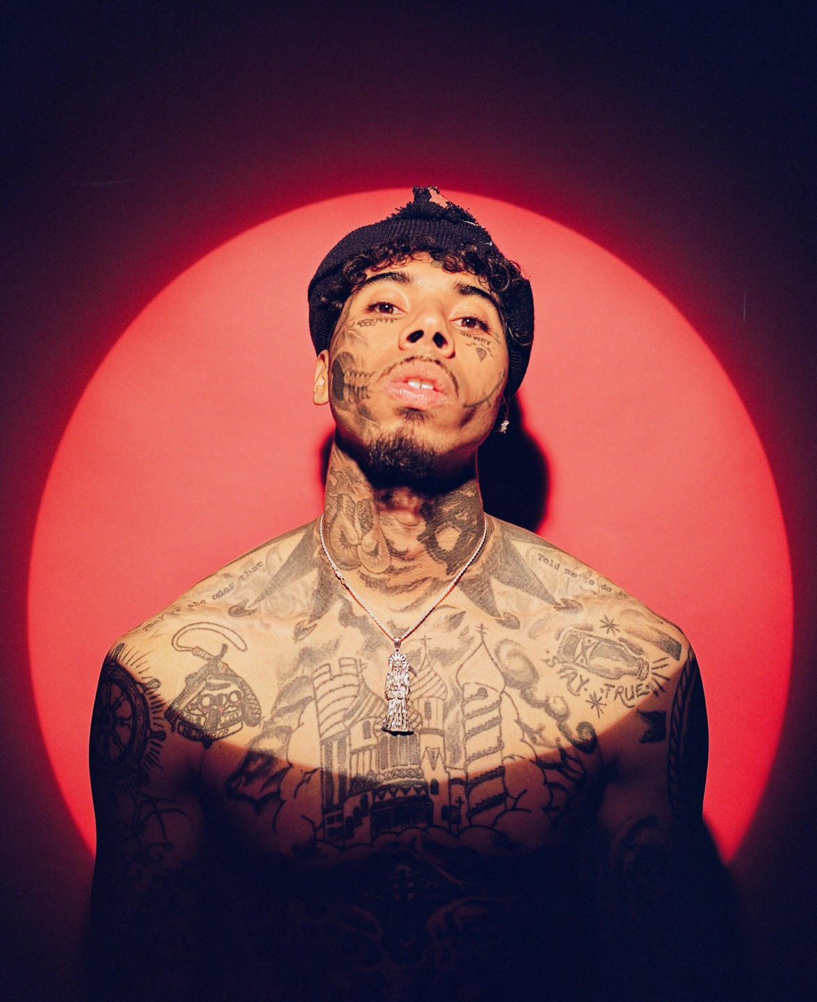 A man with many tattoos on his face, neck, and torso looks up at the camera against a red background with a circular spotlight.