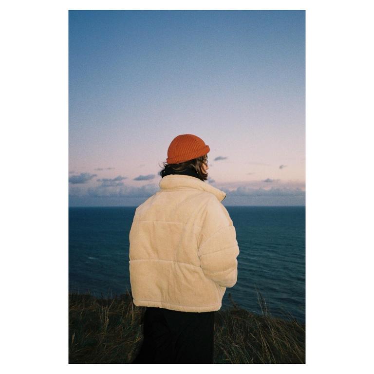 A person in a puffer jacket and beanie stands on a cliff overlooking the sea at dusk.
