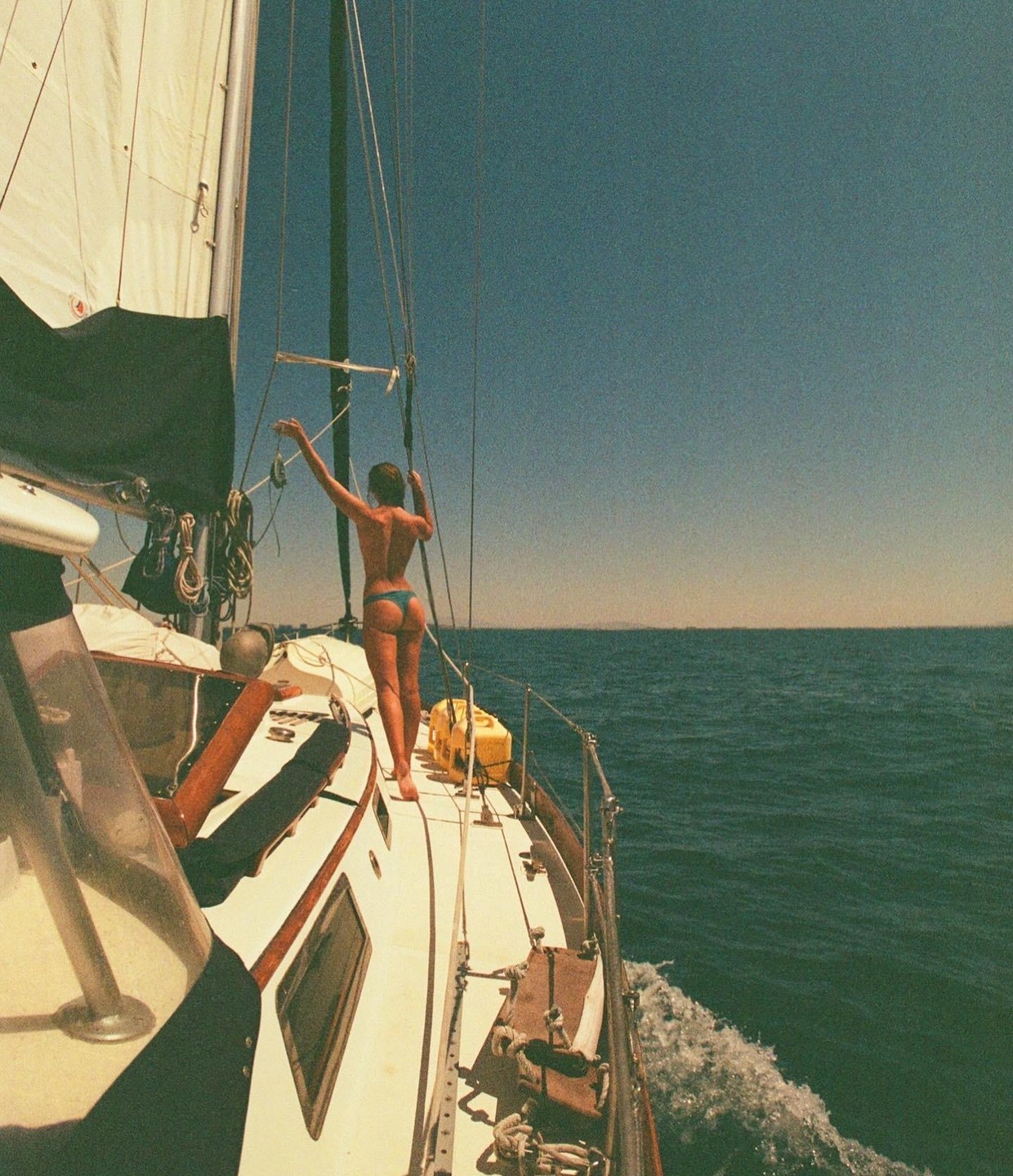 A woman in a bikini stands on the deck of a sailboat as it moves through the water.