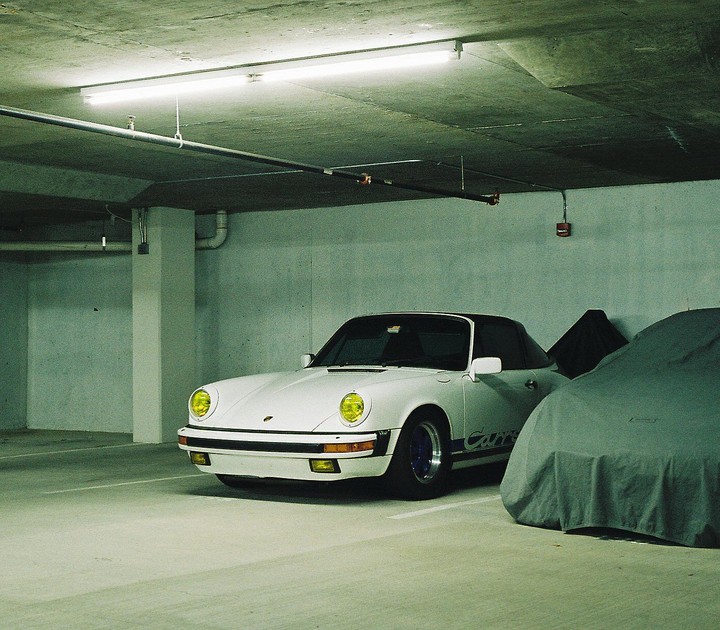 A white convertible car with yellow headlights is parked next to a car covered with a tarp in an underground parking garage.