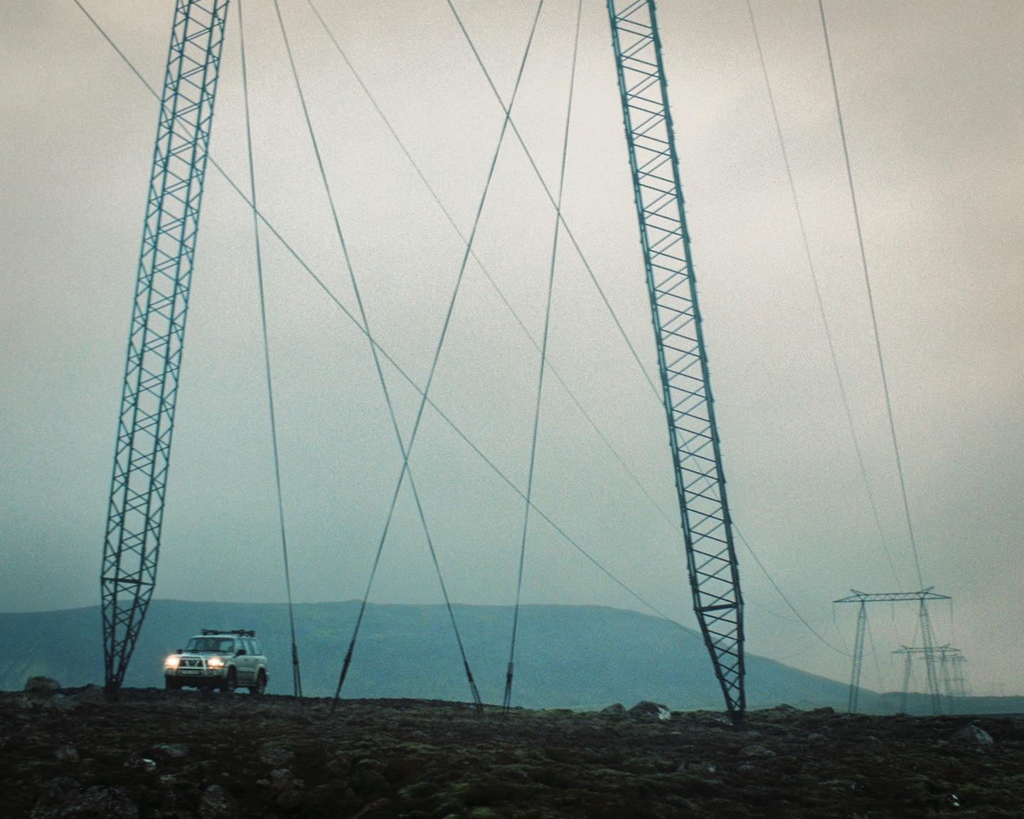 A car with its headlights on is parked on a rocky, mossy hill between two large power line towers, with more towers and mountains in the hazy background.