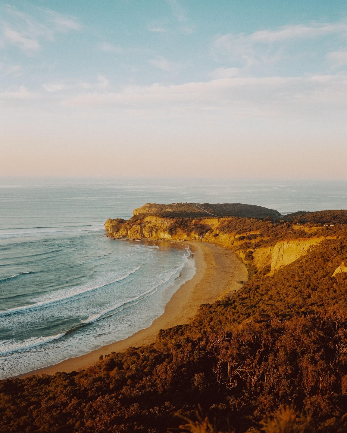 A sandy beach curves along a coastline with cliffs and green foliage under a blue sky.