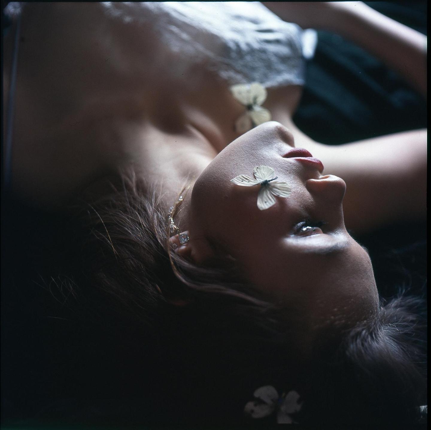 A woman lies on her back with a butterfly on her eyelid and another on her chest.