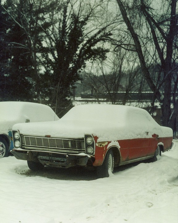 A red car is parked in the snow next to another snow-covered vehicle, with bare trees in the background.