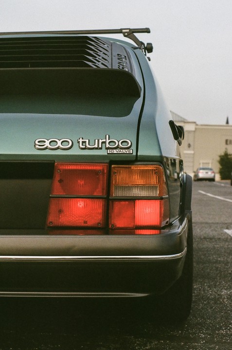 The rear of a green 900 Turbo car is parked on asphalt with its taillights visible.