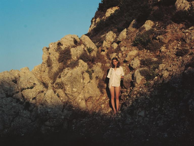 A woman stands on a rocky hillside with a clear sky above.