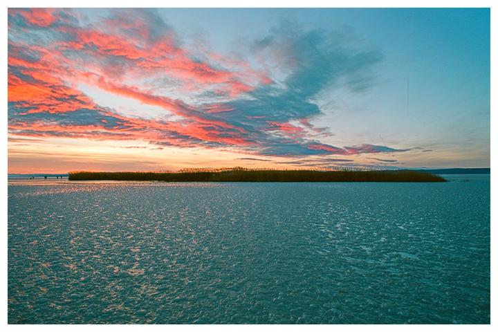 A frozen lake with a strip of reeds in the distance is illuminated by the vibrant colors of a sunset sky.