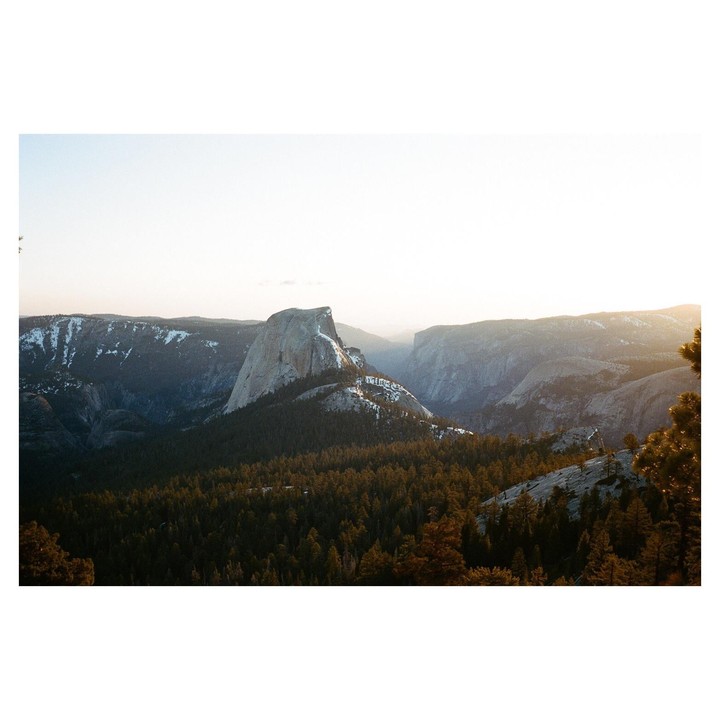 A mountain range with a prominent rock formation is covered in trees and patches of snow under a bright sky.