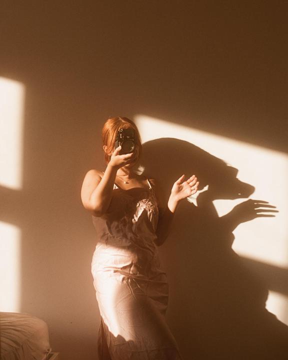 A woman in a dress holds a camera to her face, casting shadows on a wall.
