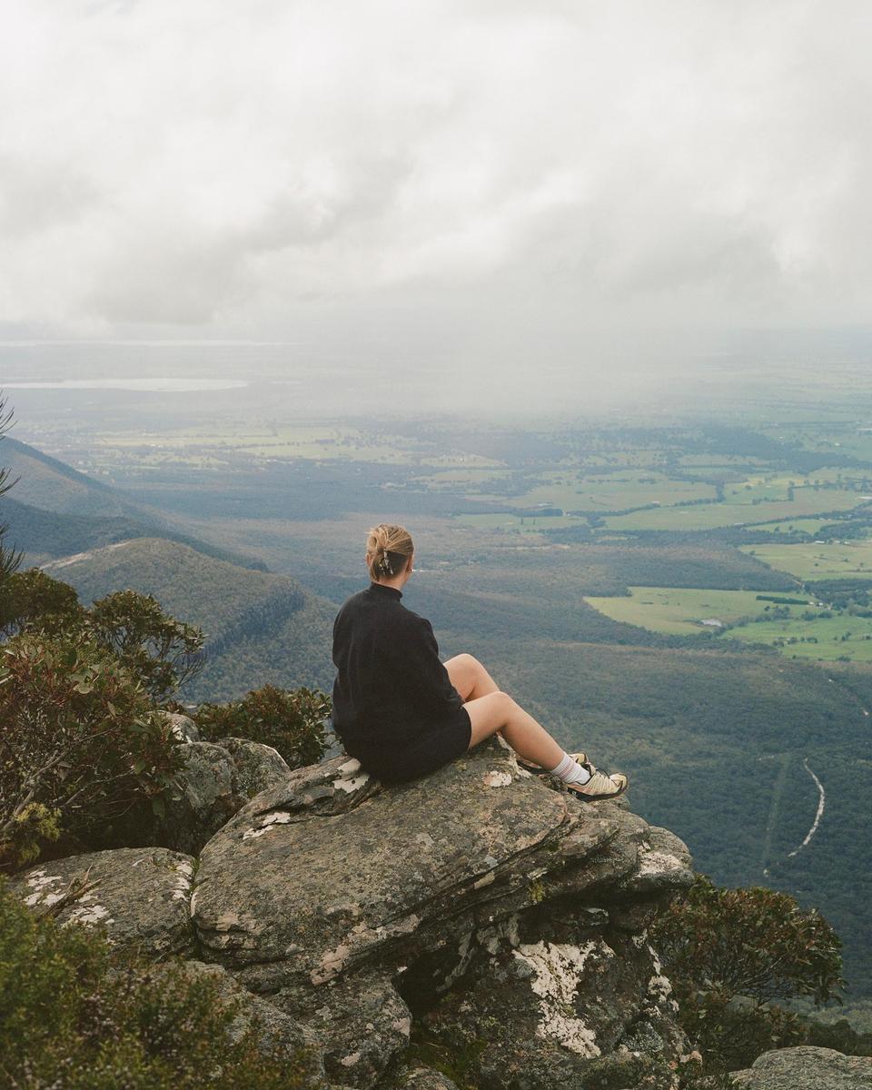 A woman sits on a rocky outcrop overlooking a vast landscape of forests and fields under a cloudy sky.