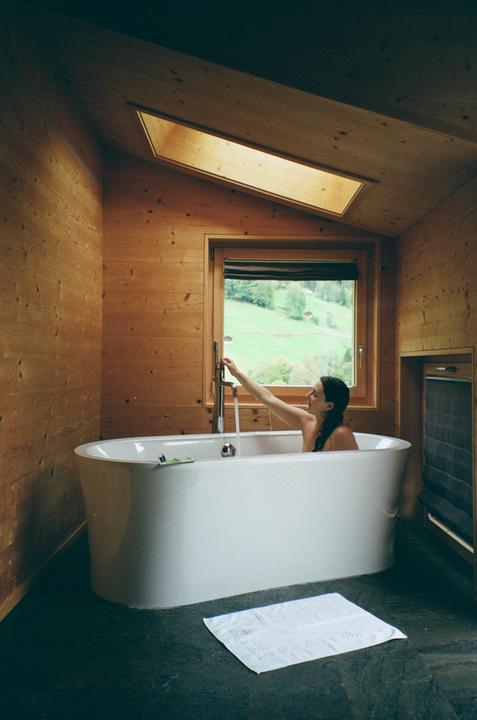 A woman sits in a bathtub filling with water in a wood-paneled bathroom with a view of a green hillside.
