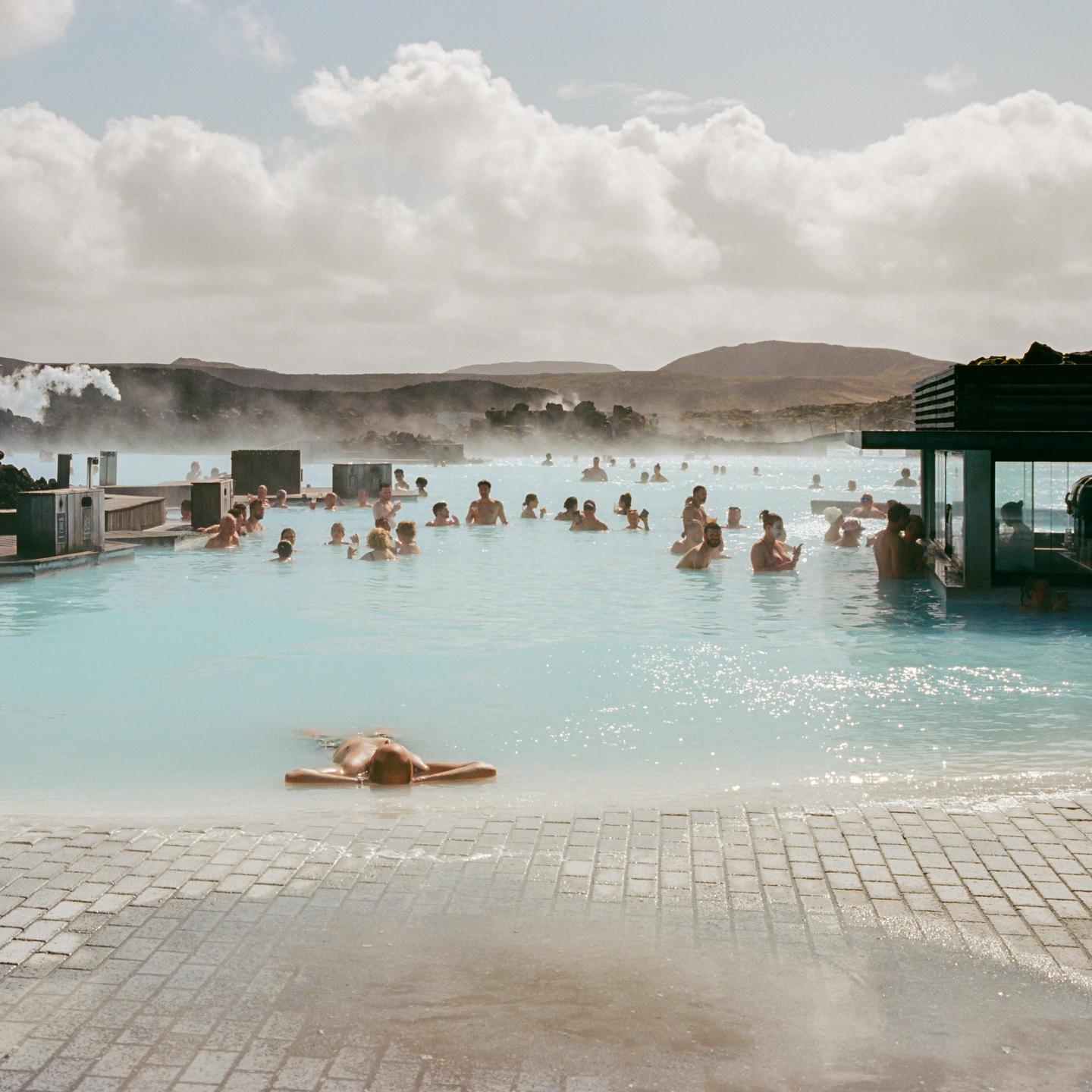 Many people are swimming in a light blue geothermal pool with steam rising from the water and mountains in the background.