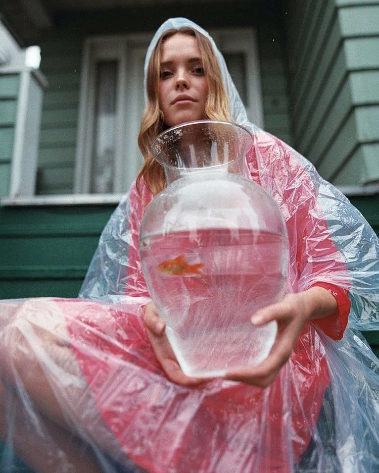 A woman wearing a clear plastic raincoat holds a large glass vase with a goldfish inside.