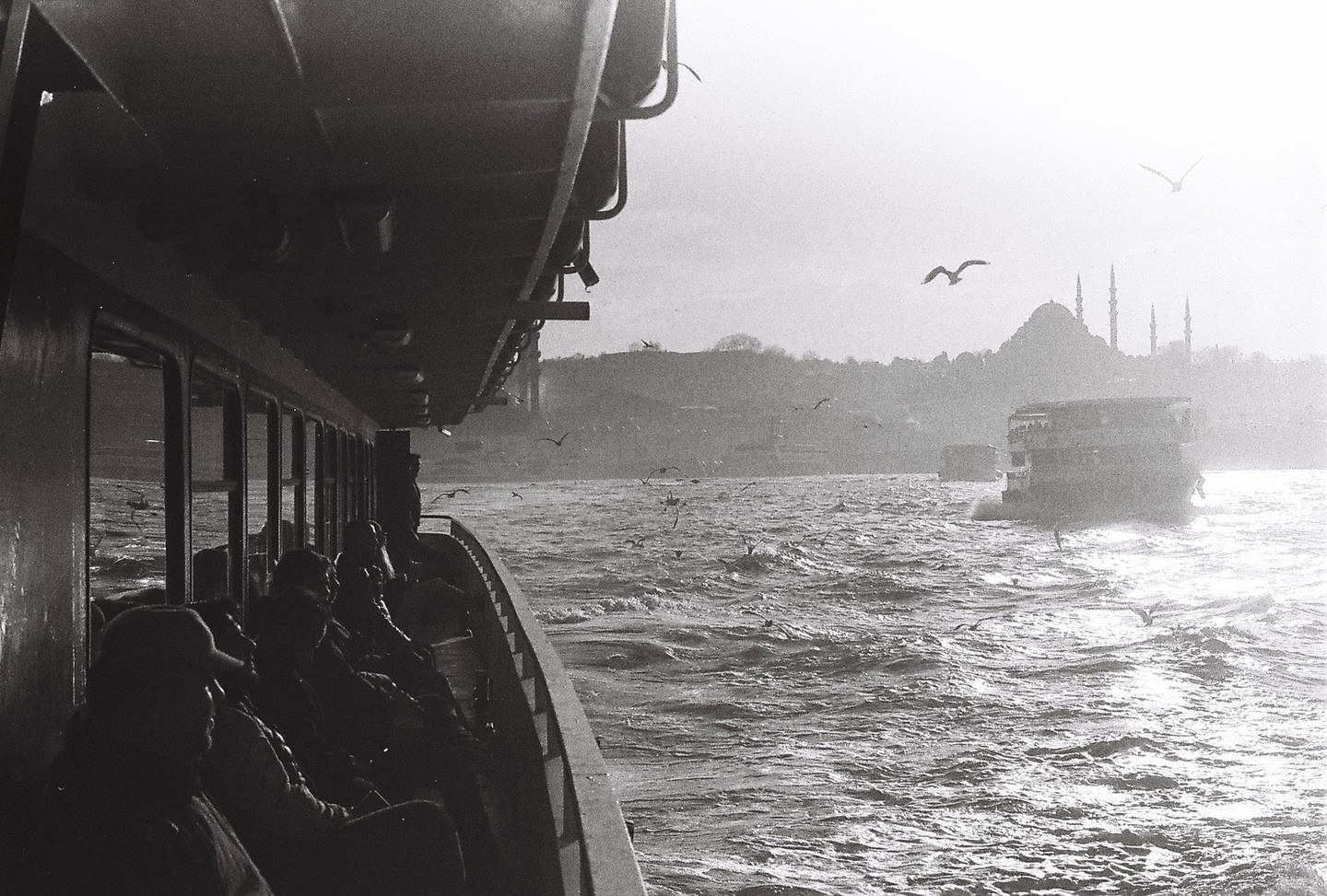 People sit on a ferry boat as it travels across choppy water with a mosque visible in the distance.