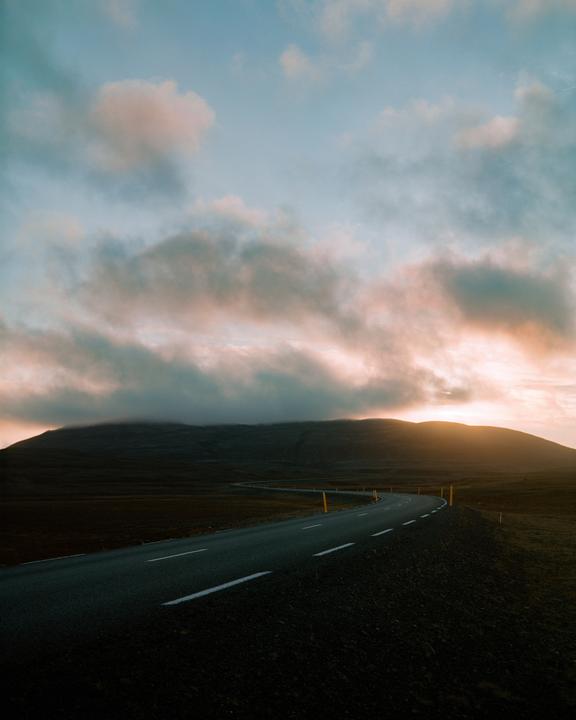 A winding road leads towards a sunset over a mountain range.