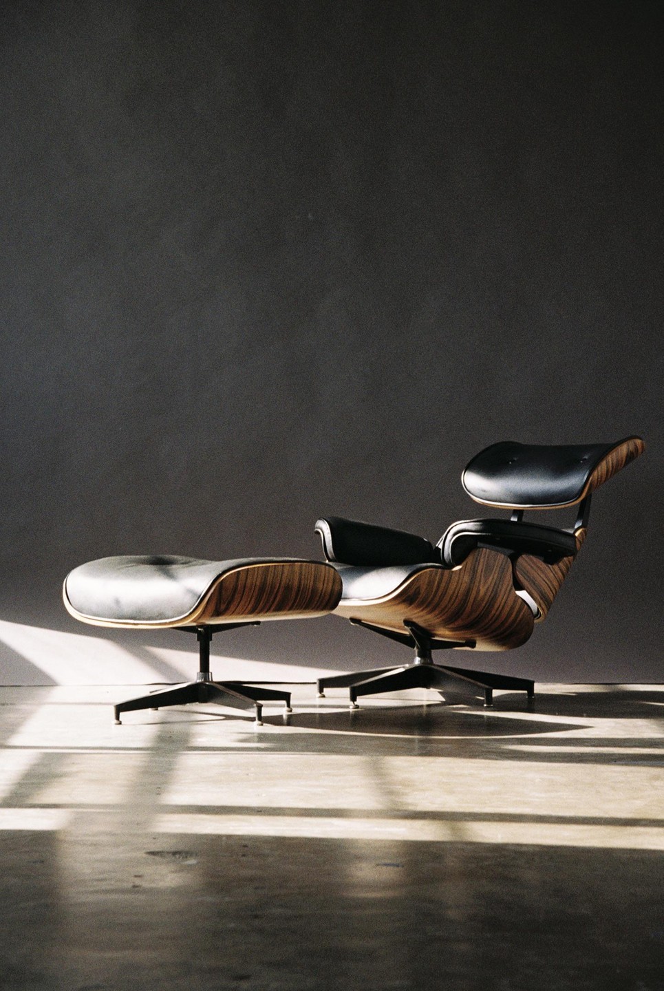 A black leather and wood lounge chair with an ottoman sits on a concrete floor with shadows cast across it.