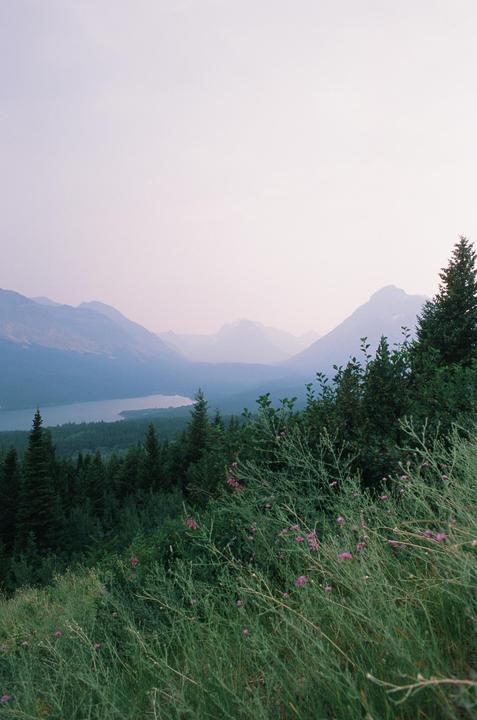 A hazy mountain landscape shows a lake surrounded by forest and mountains under a pale sky.
