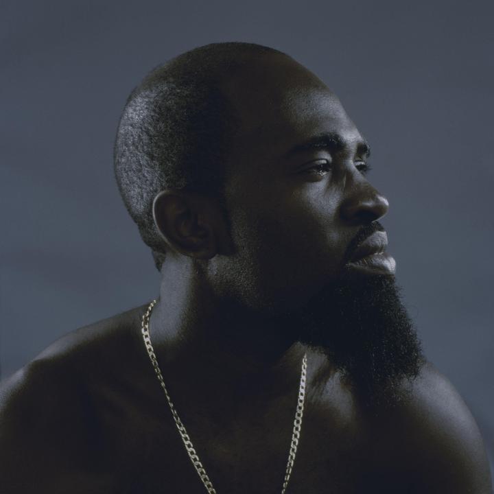 A man with a beard and a gold chain looks up and to the right.