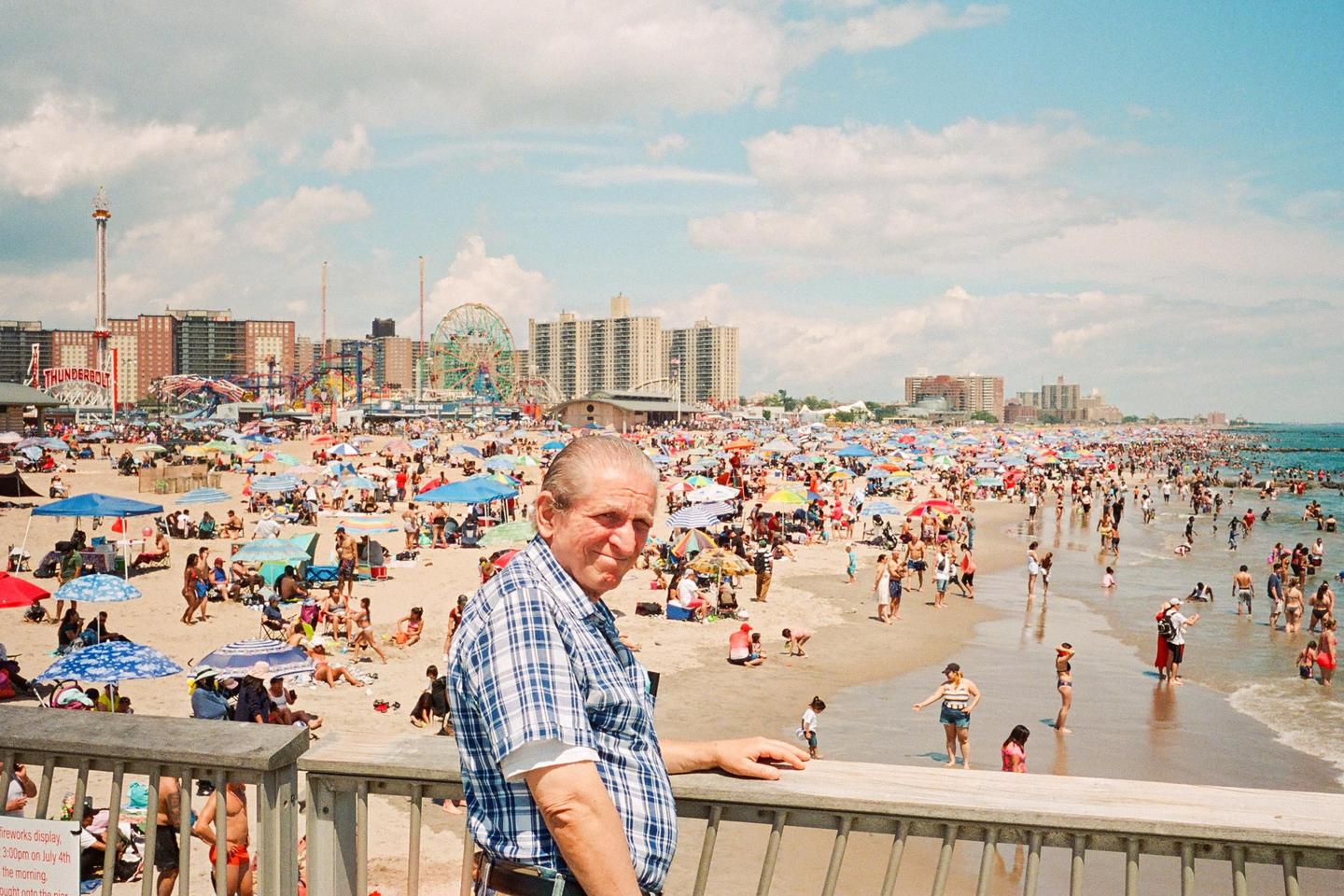 A man in a plaid shirt stands on a pier overlooking a crowded beach with an amusement park in the background.