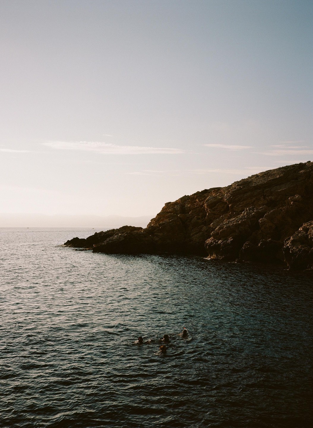 Four people swim in the ocean next to a rocky cliff.