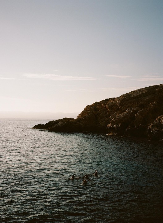 Four people swim in the ocean next to a rocky cliff.
